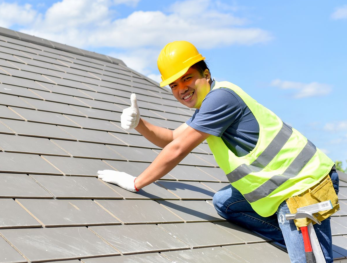 Roofer on a gray tile roof gives a thumbs up; wearing a yellow helmet, safety vest, and holding a tool.