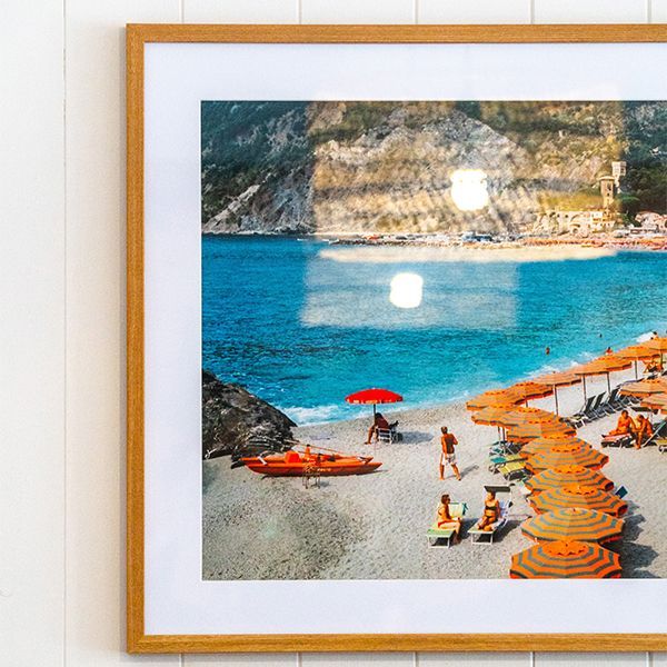 Framed beach painting with turquoise water, rocky cliffs, orange loungers, and a red umbrella on the shore