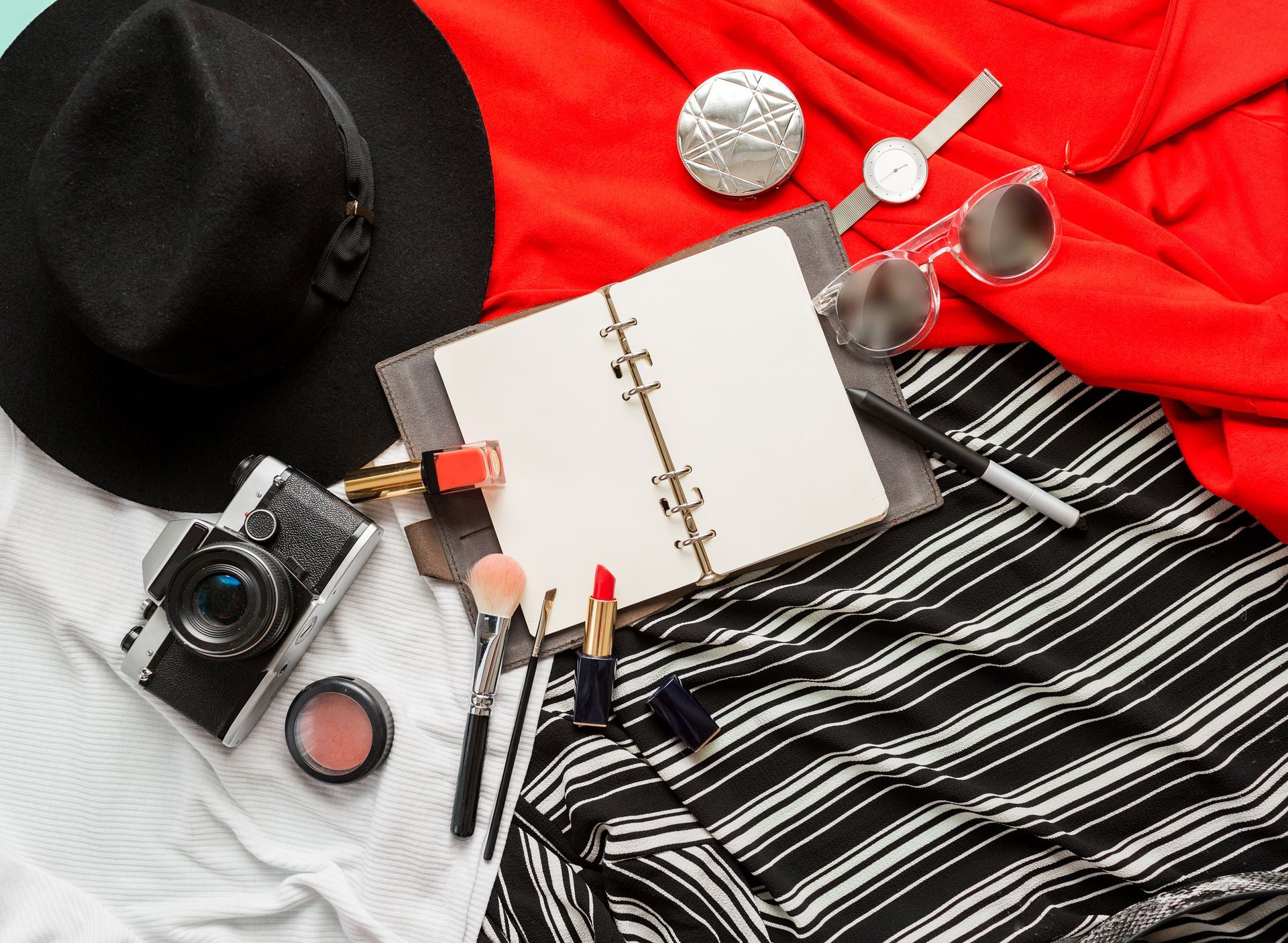 A hat , camera , sunglasses , lipstick , brushes and a notebook are on a table.