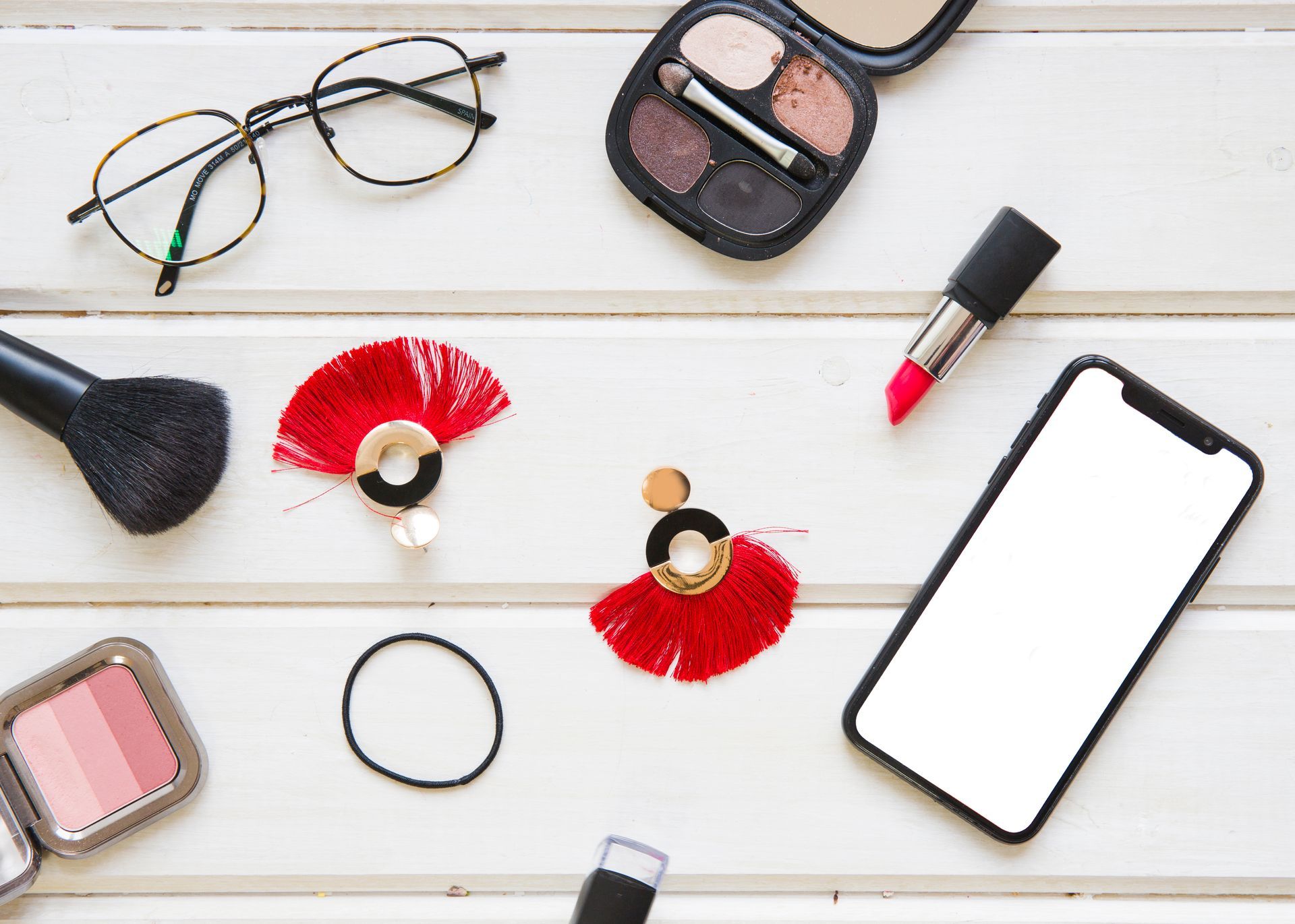 A white wooden table topped with cosmetics , a cell phone , and a pair of glasses.