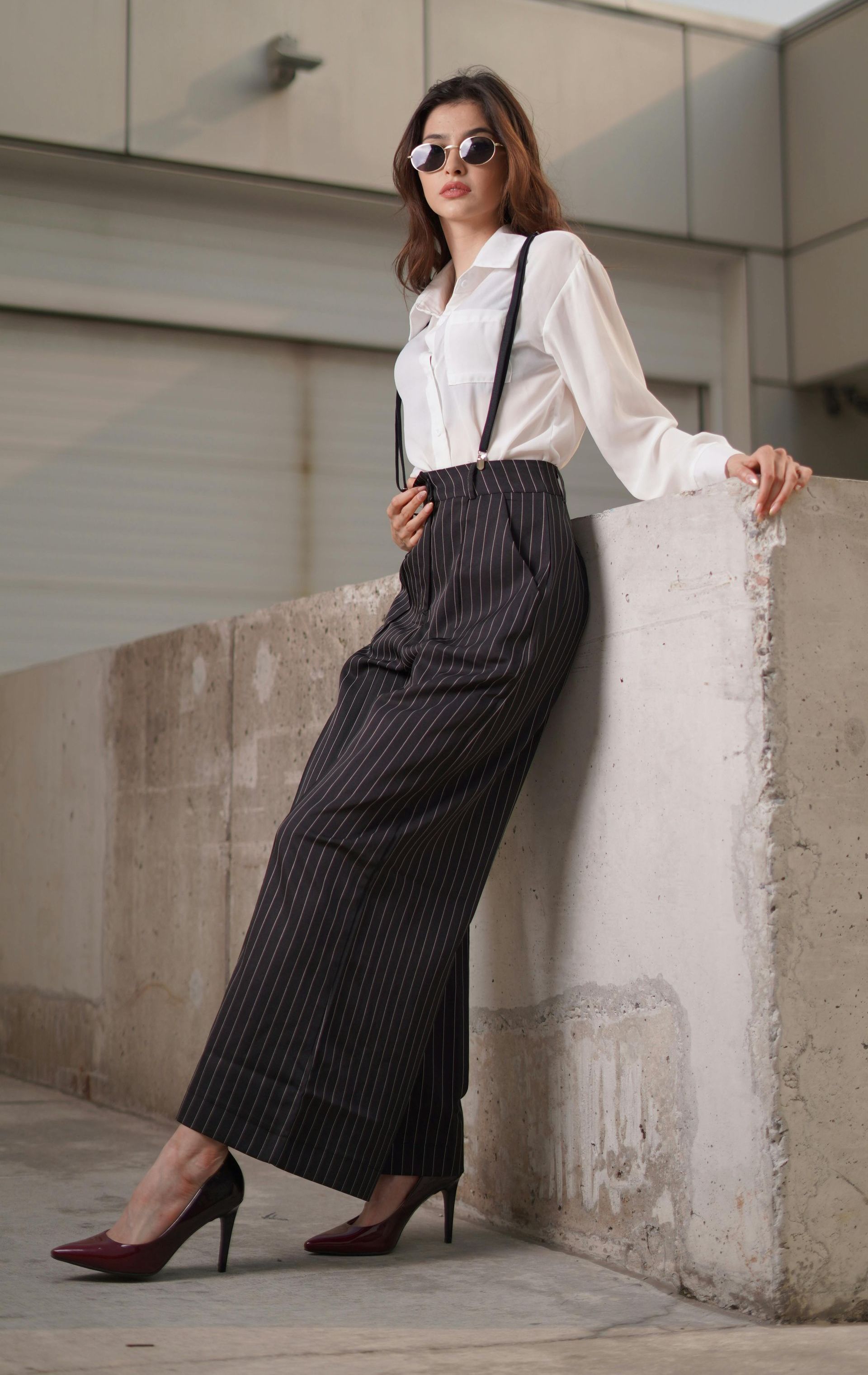 A woman wearing a white shirt and suspenders is leaning against a wall.