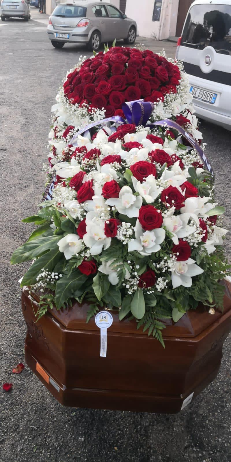 Una bara ricoperta da una composizione floreale di fiori rossi e bianchi. Sullo sfondo si vedono delle auto.