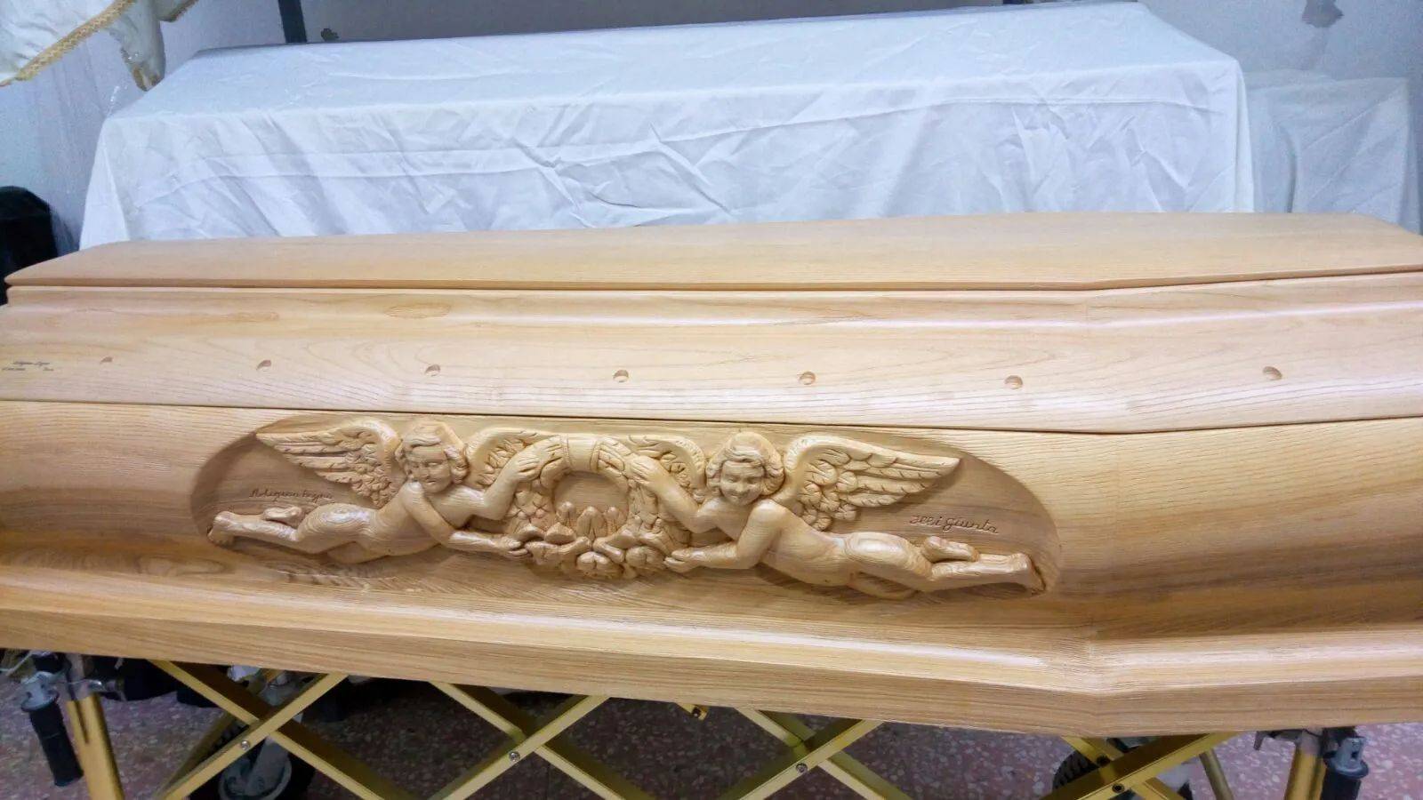 Bara di legno con cherubini intagliati, appoggiata su un supporto giallo. Sullo sfondo, un lenzuolo bianco su un tavolo.