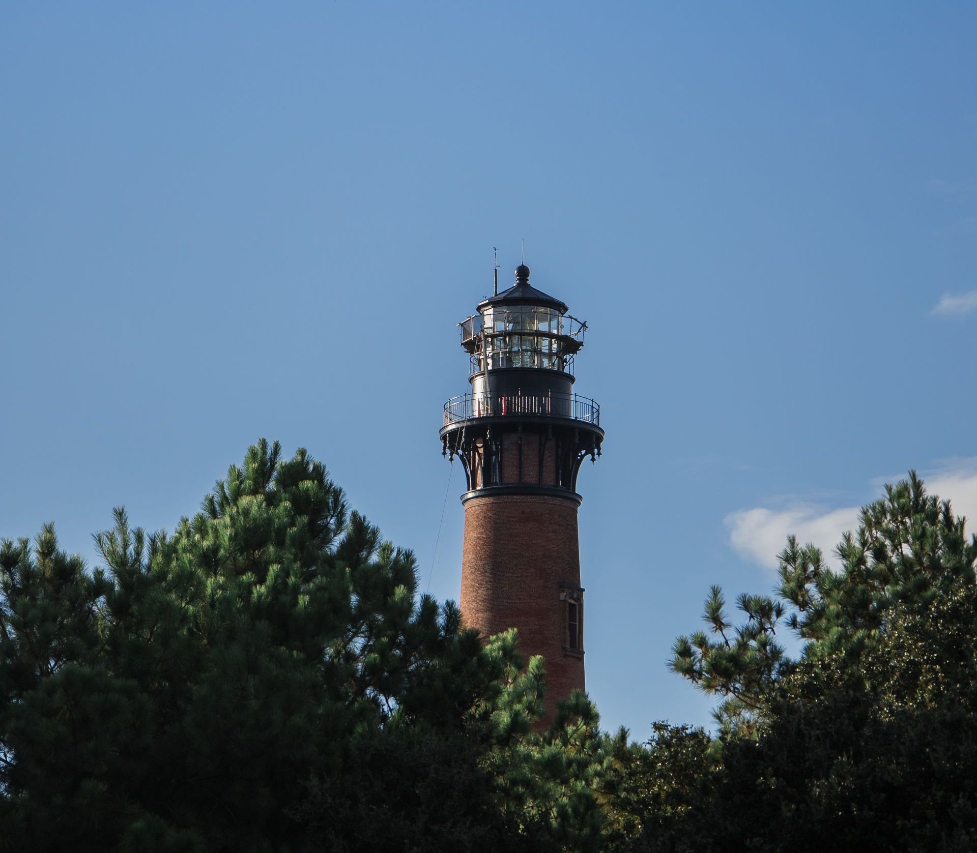 A tall brick lighthouse is surrounded by trees and a blue sky