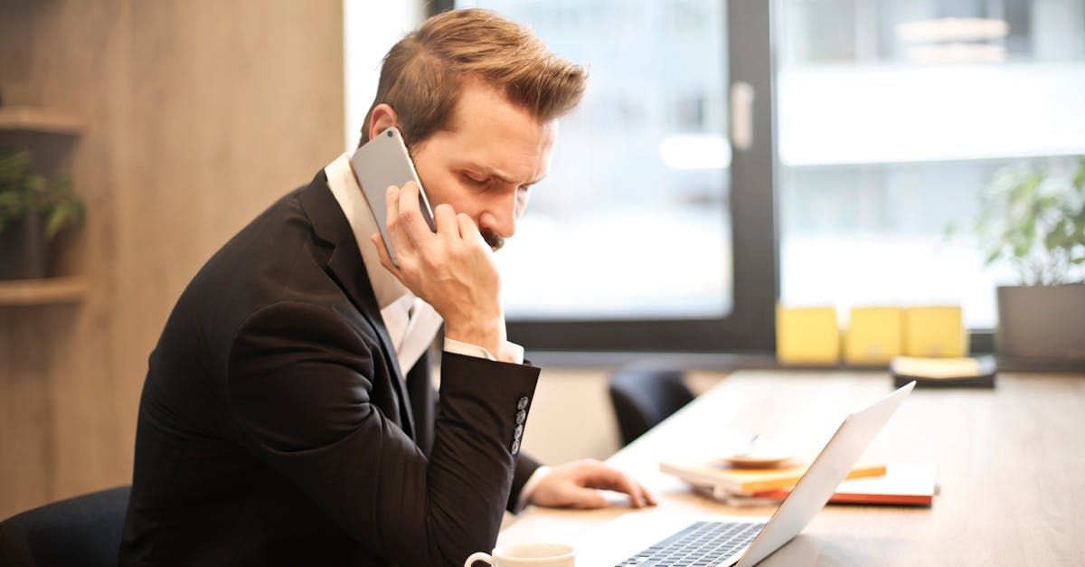 A man in a suit is talking on a cell phone while using a laptop computer.