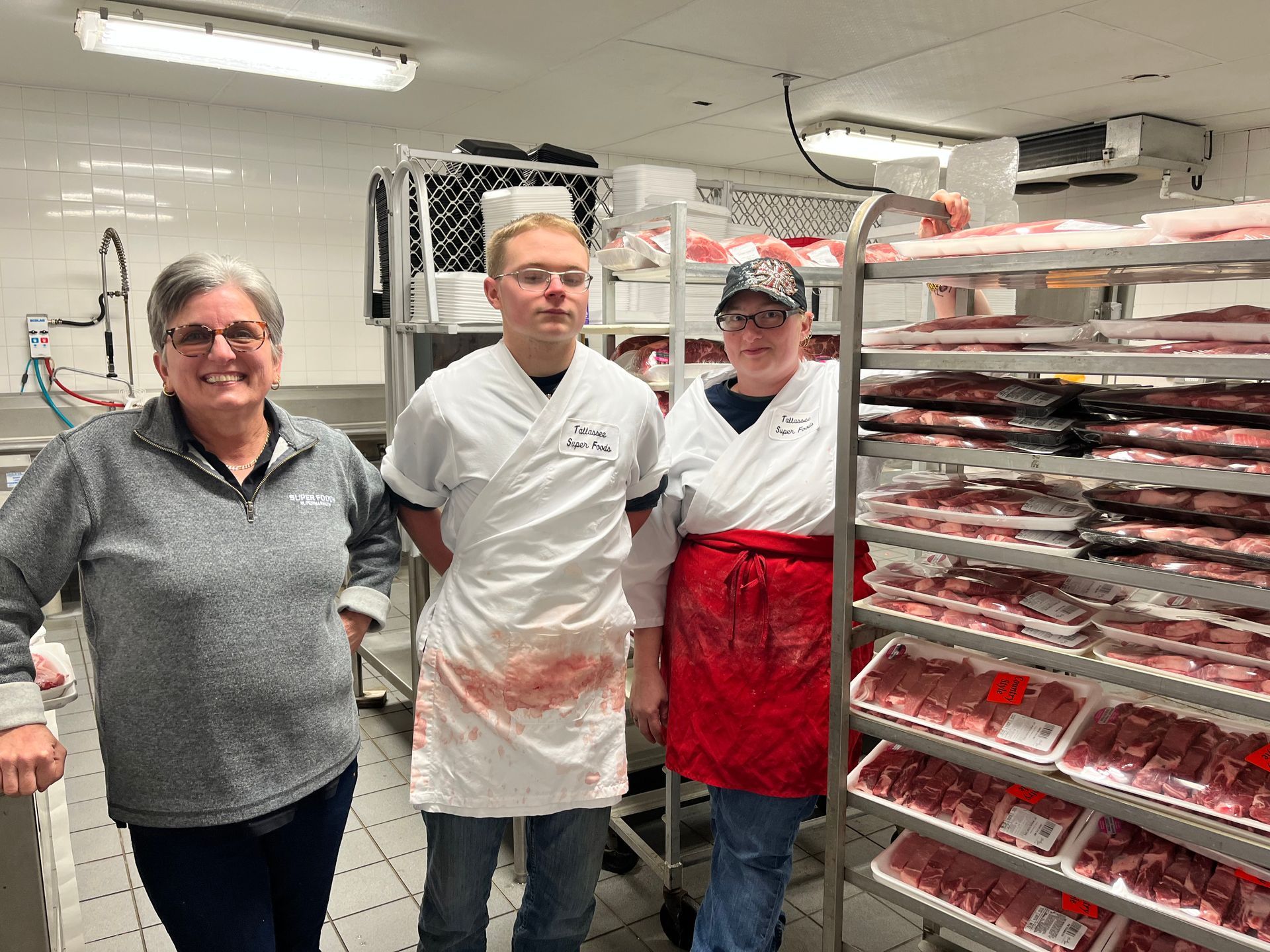 Three people in a meat processing room stand near shelves of packaged meat. Two wear aprons.