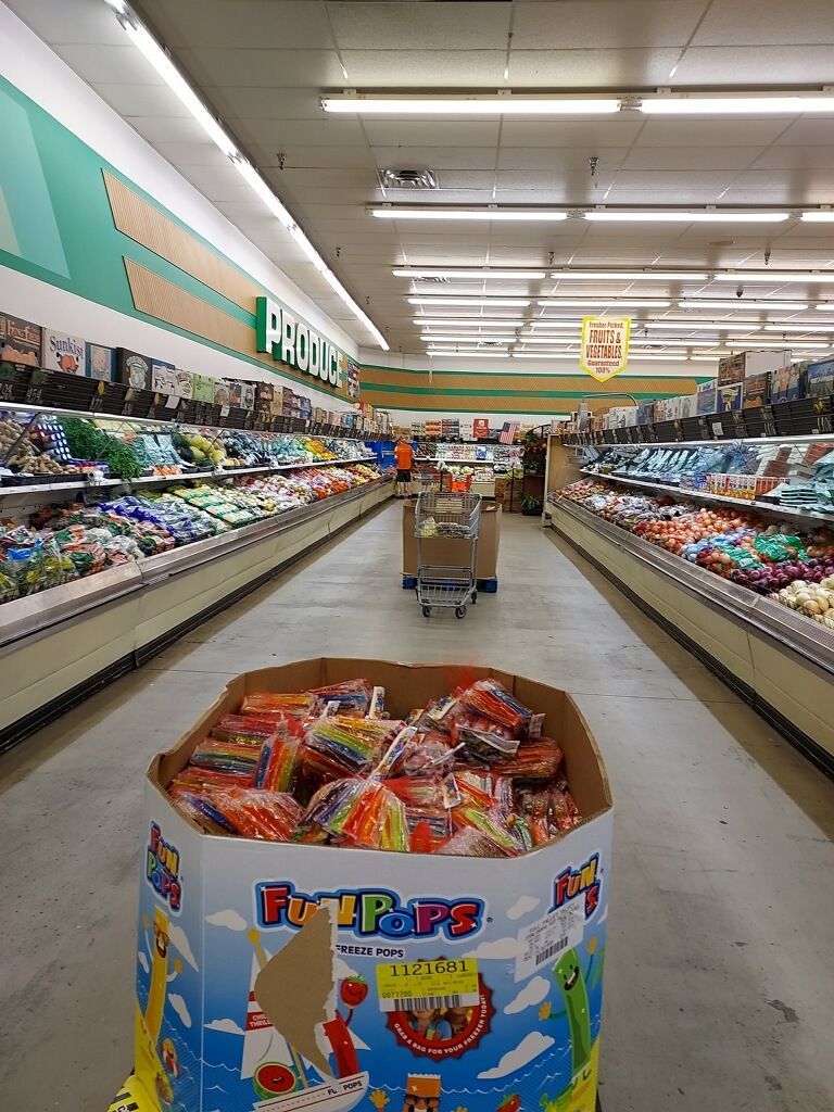 Grocery store aisle with candy display in foreground, produce and a shopping cart in the background.