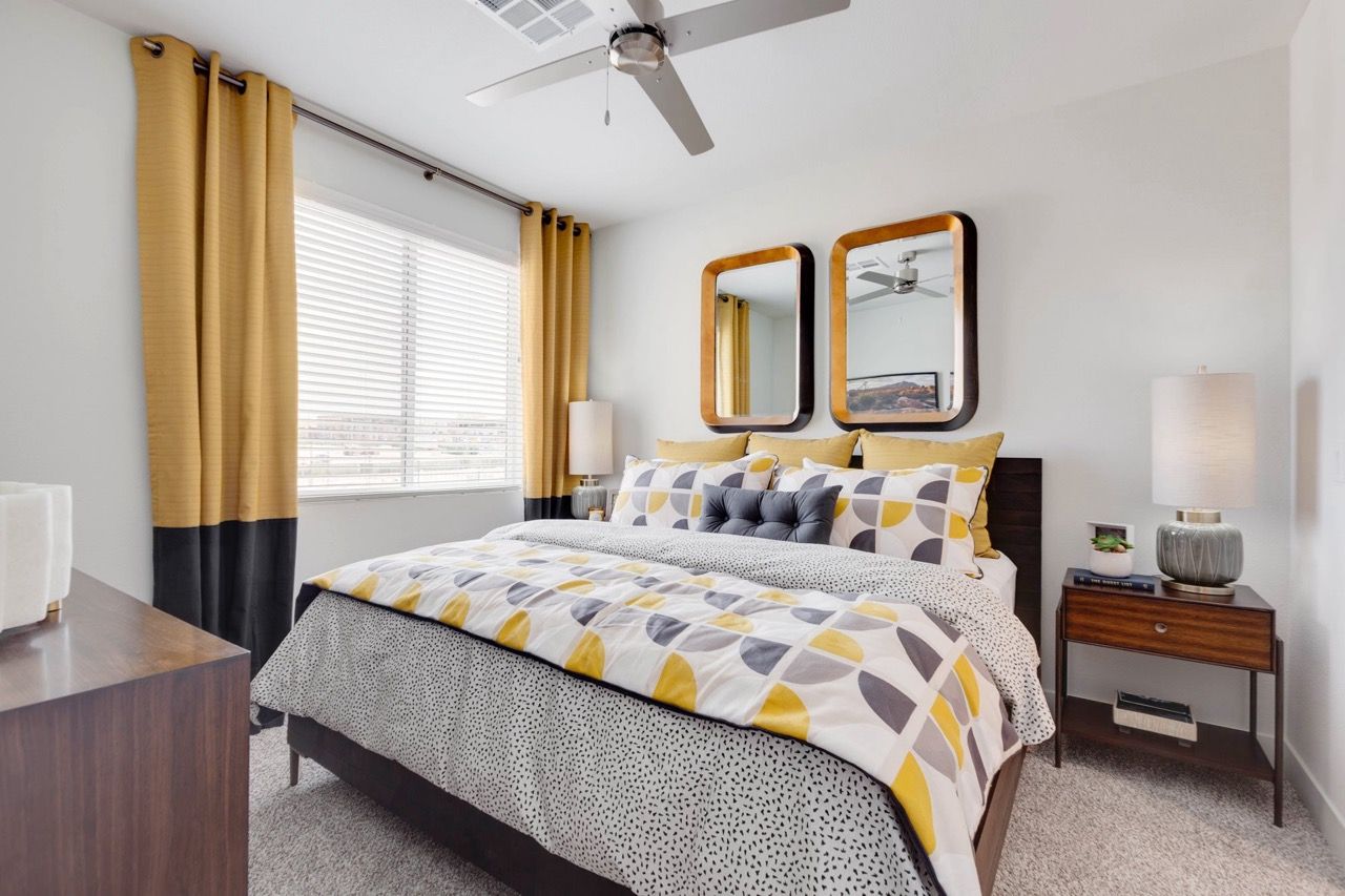 Apartment bedroom with large window, mustard curtains, patterned bedding, and two square mirrors above the headboard.