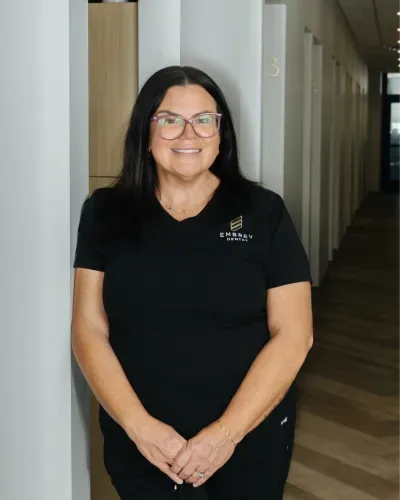 Woman in black polo shirt smiling in a hallway, standing with hands clasped.