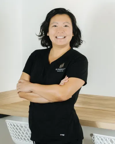 Smiling person in black scrubs stands with arms crossed beside a wooden table.