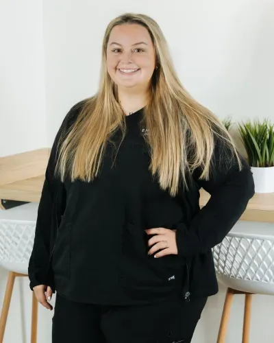 Woman in black outfit standing and smiling indoors beside a wooden table with a potted plant.