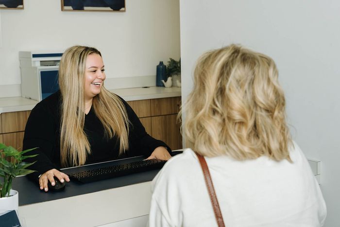 Receptionist smiling at a patient at the front desk. Blonde hair, black top, office setting.