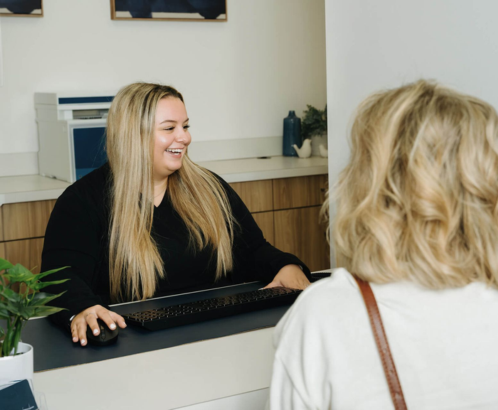 Receptionist smiling at a patient at the front desk. Blonde hair, black top, office setting.