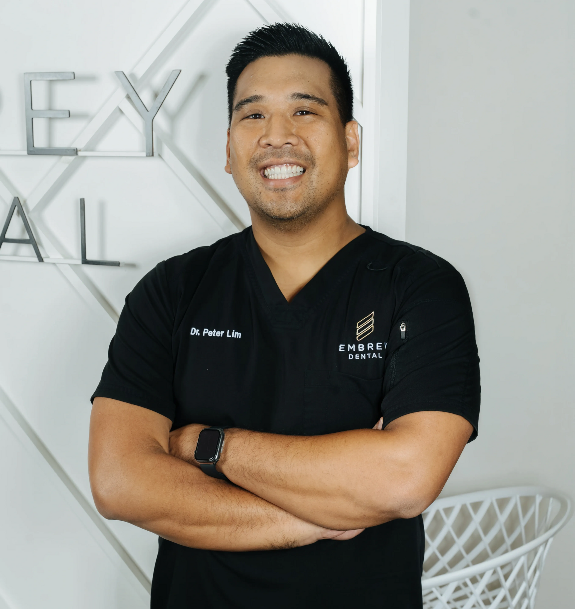 Smiling person in black polo with arms crossed, standing in a bright office setting.
