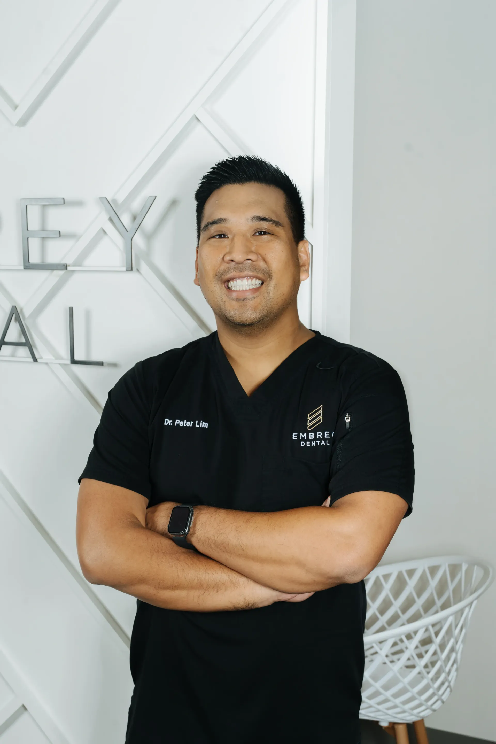 Smiling person in black scrubs standing with arms crossed against a white wall with geometric decor.