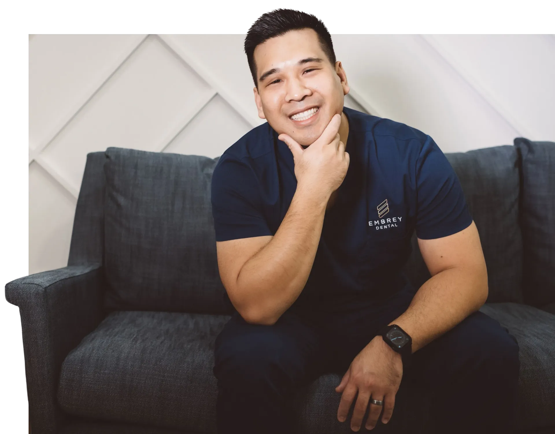Doctor smiling, seated on a gray couch, chin in hand. Navy blue shirt. Bright white background.