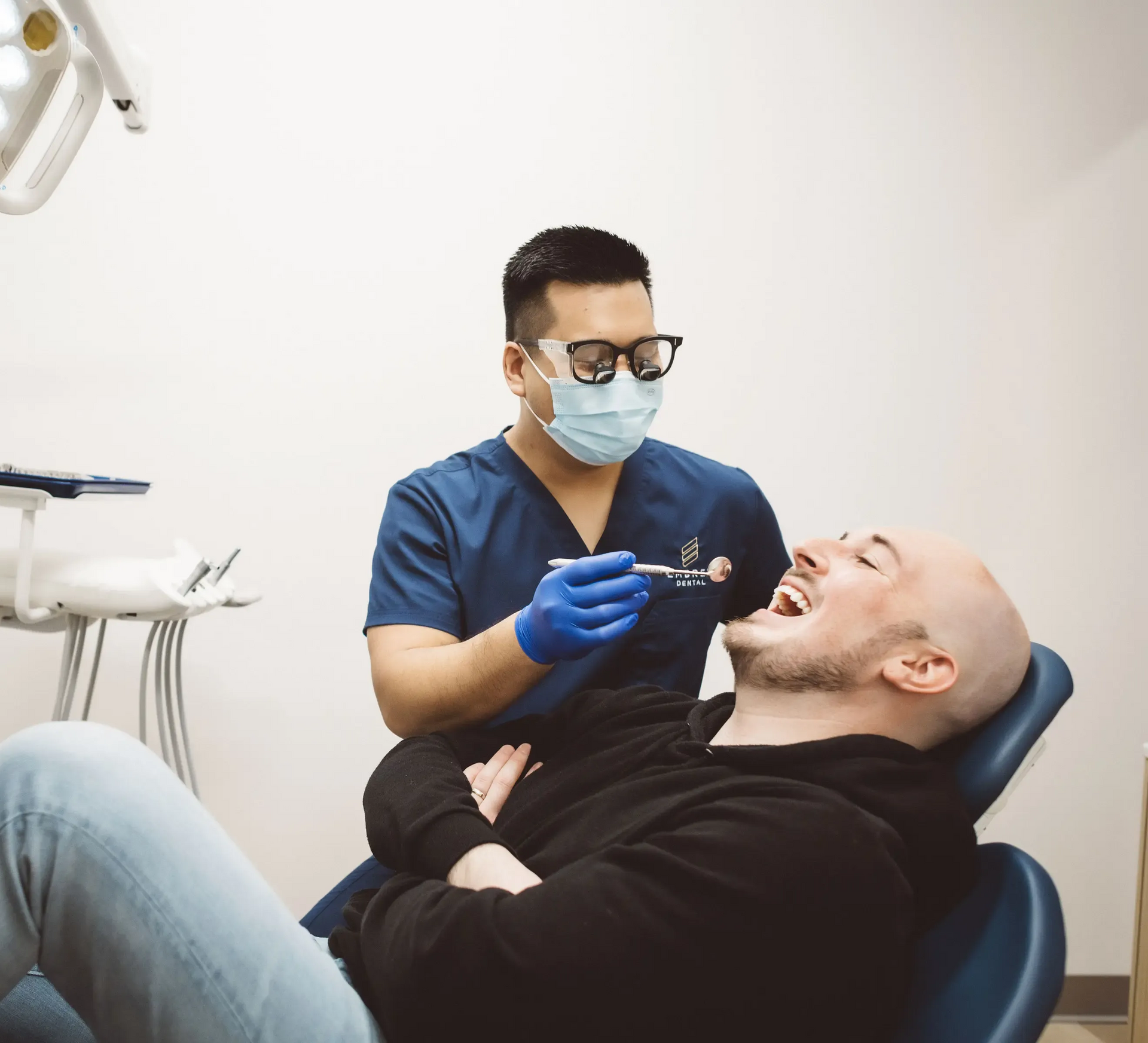 A dentist in scrubs and a mask examines a patient's teeth in a clinical setting.
