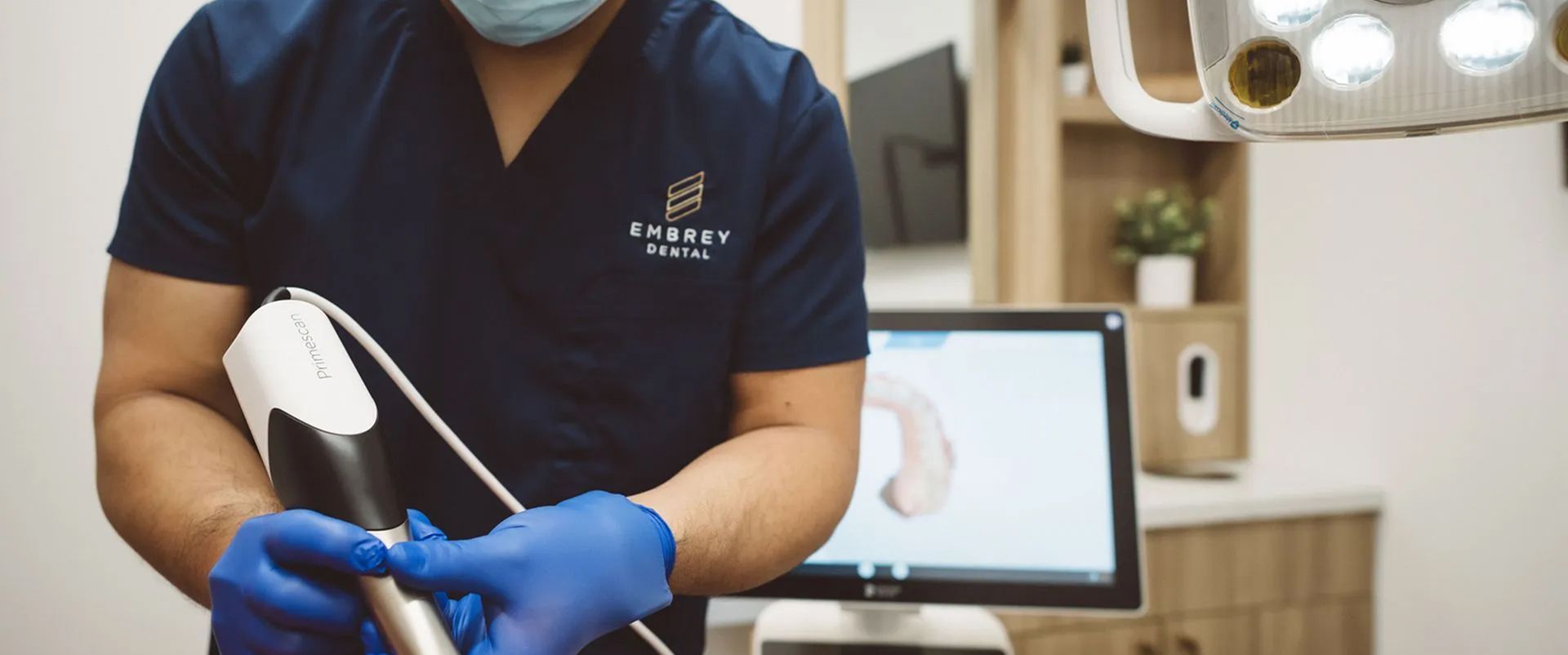 Gloved technician holding dental tools beside a patient chair, with a monitor in the background