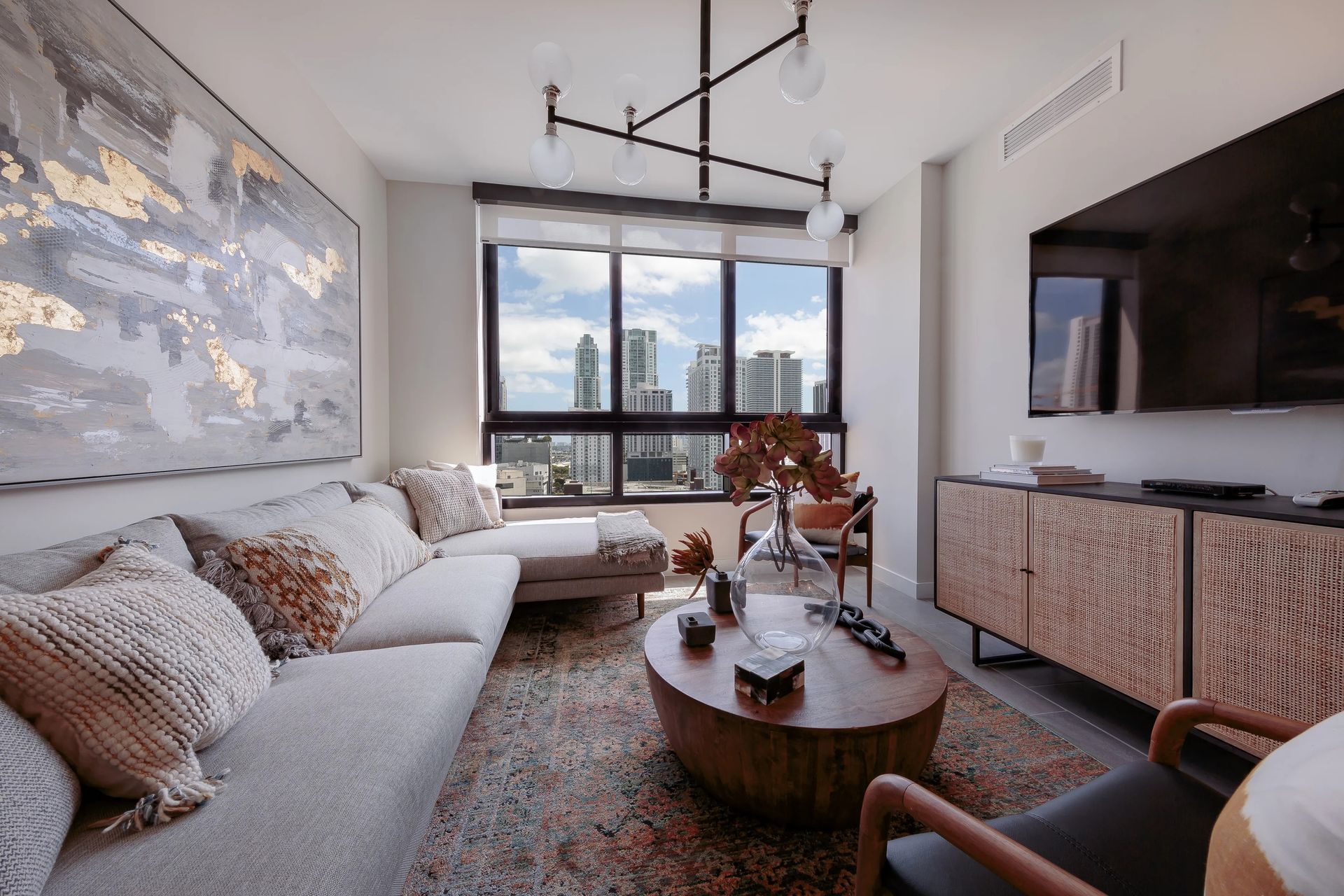 Cozy living room with light-colored sofa, wooden furniture, artwork, and city view from the window.