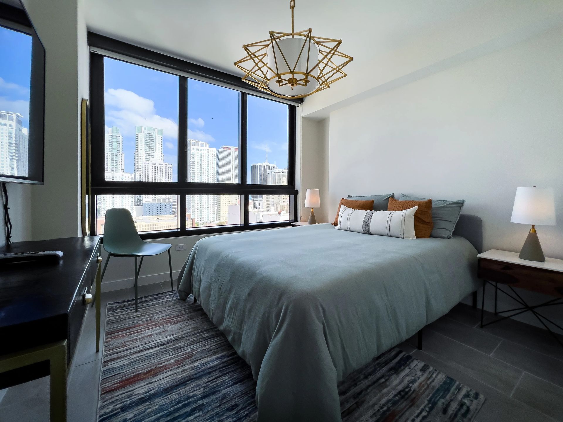 Bedroom with a bed, rug, chair, and city view through large windows.