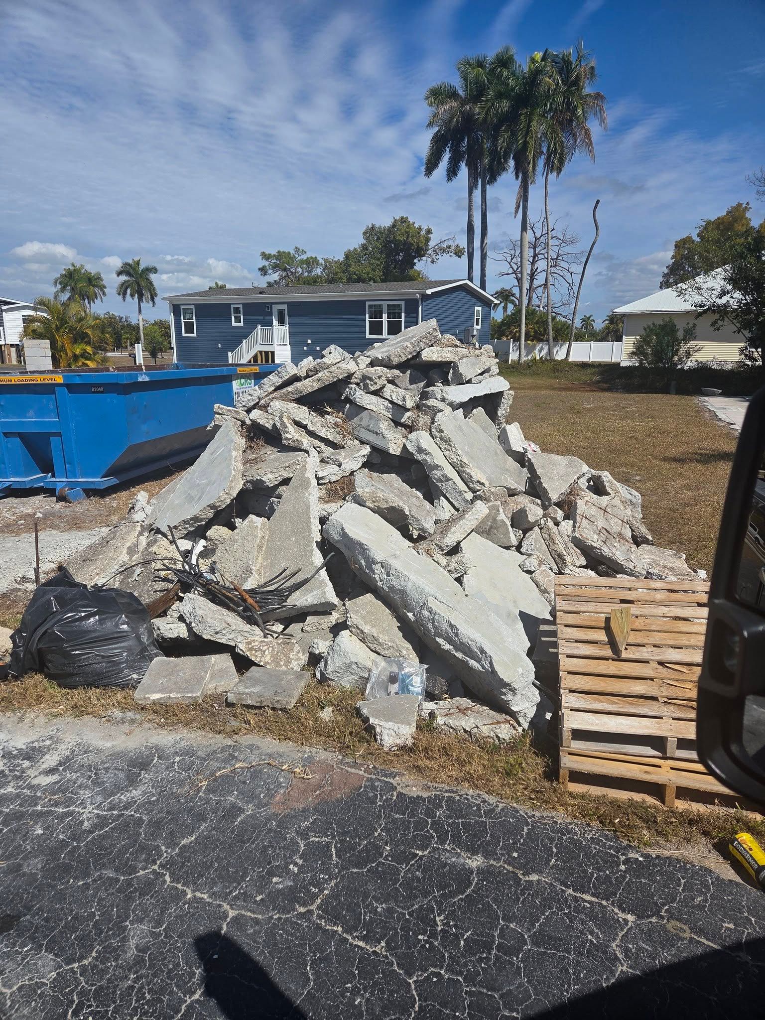 Pile of broken concrete beside a blue dumpster and wooden pallets in a sunny outdoor lot