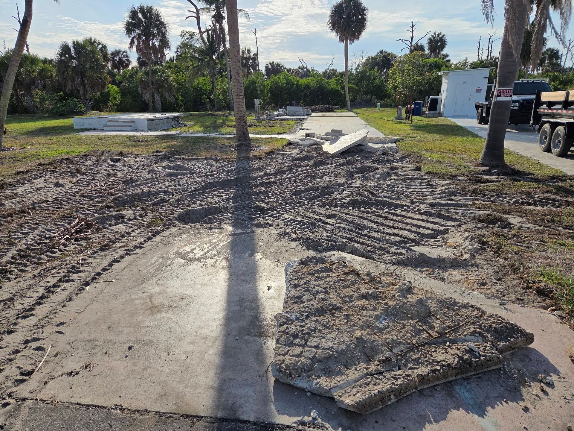 Damaged concrete walkway in a yard, with broken slabs and muddy ground under a palm tree.
