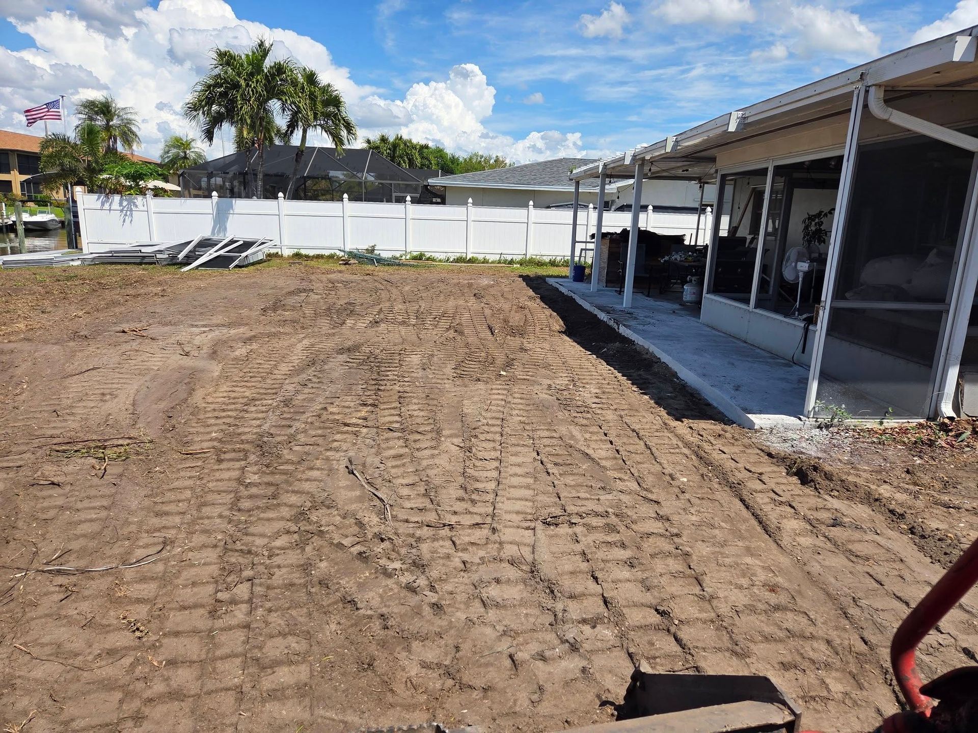 Dirt-covered lot beside a covered patio and white fence under a sunny sky