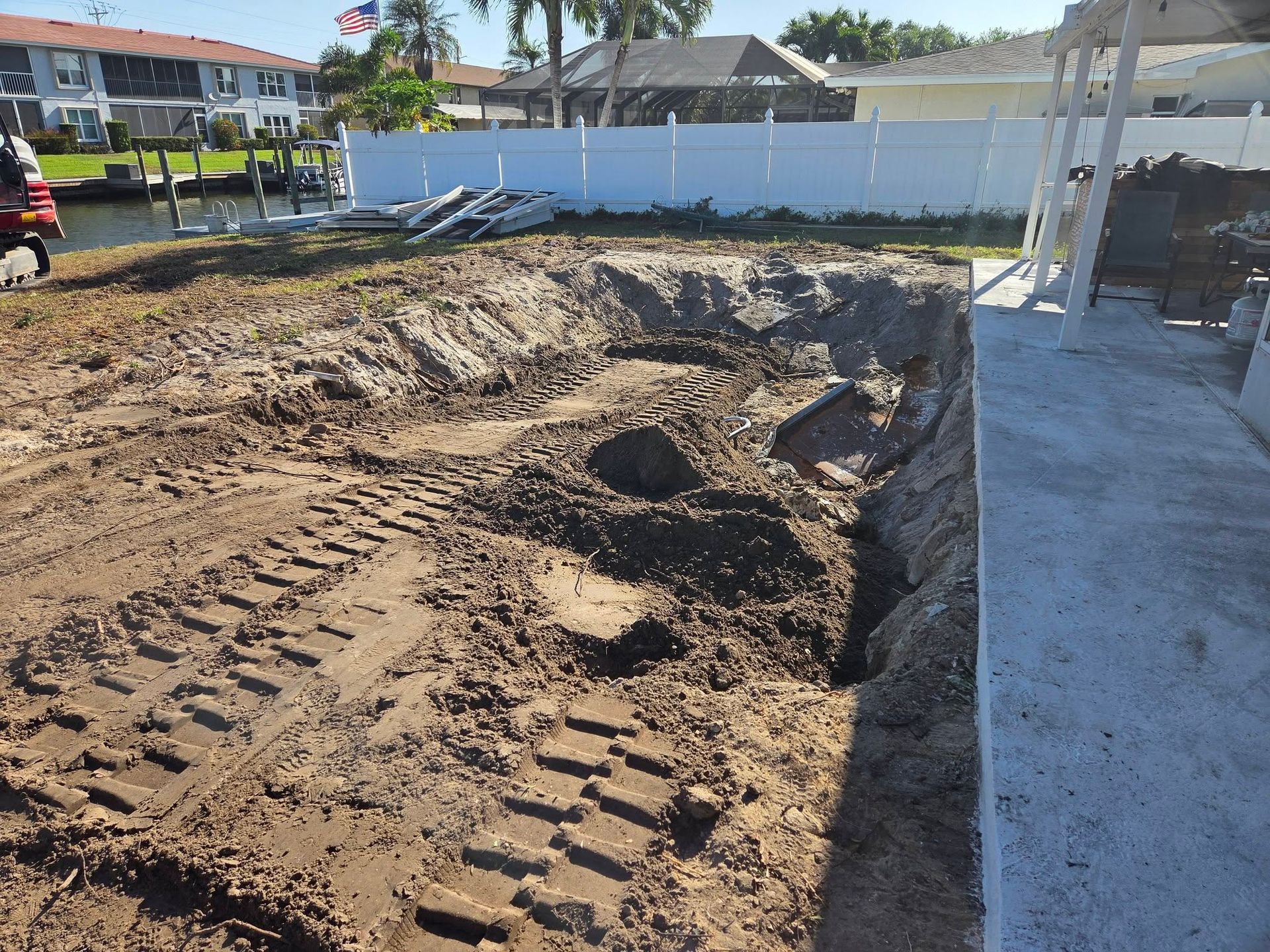 Construction site with excavated dirt beside a concrete patio and white fence under a sunny sky