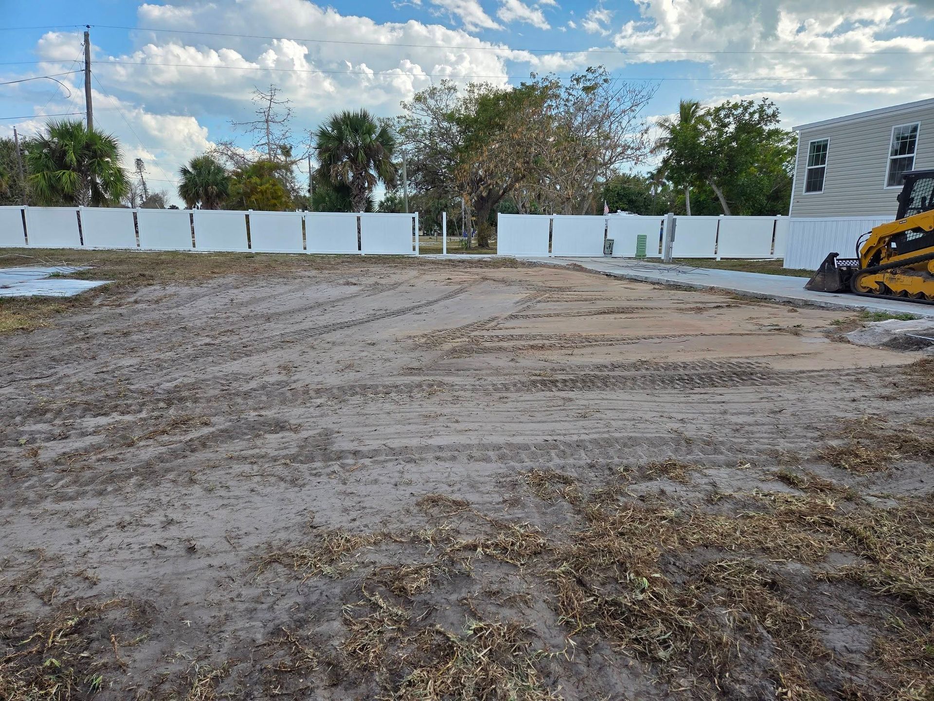 Wide dirt lot with tire tracks, white fence, trees, cloudy sky, and a yellow excavator at the right edge