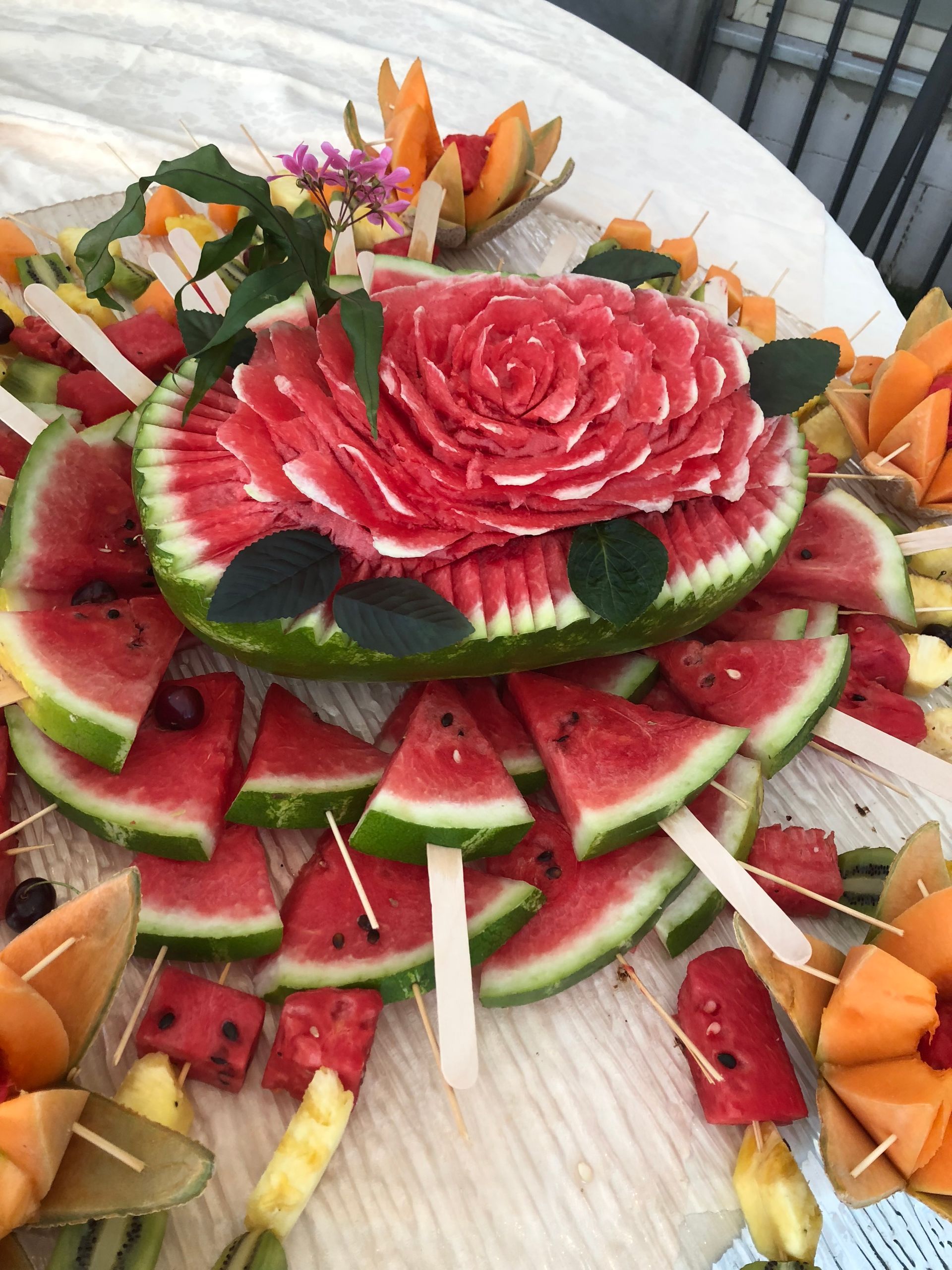 Piatto di anguria e frutta con centrotavola a forma di rosa intagliata e spiedini di melone.