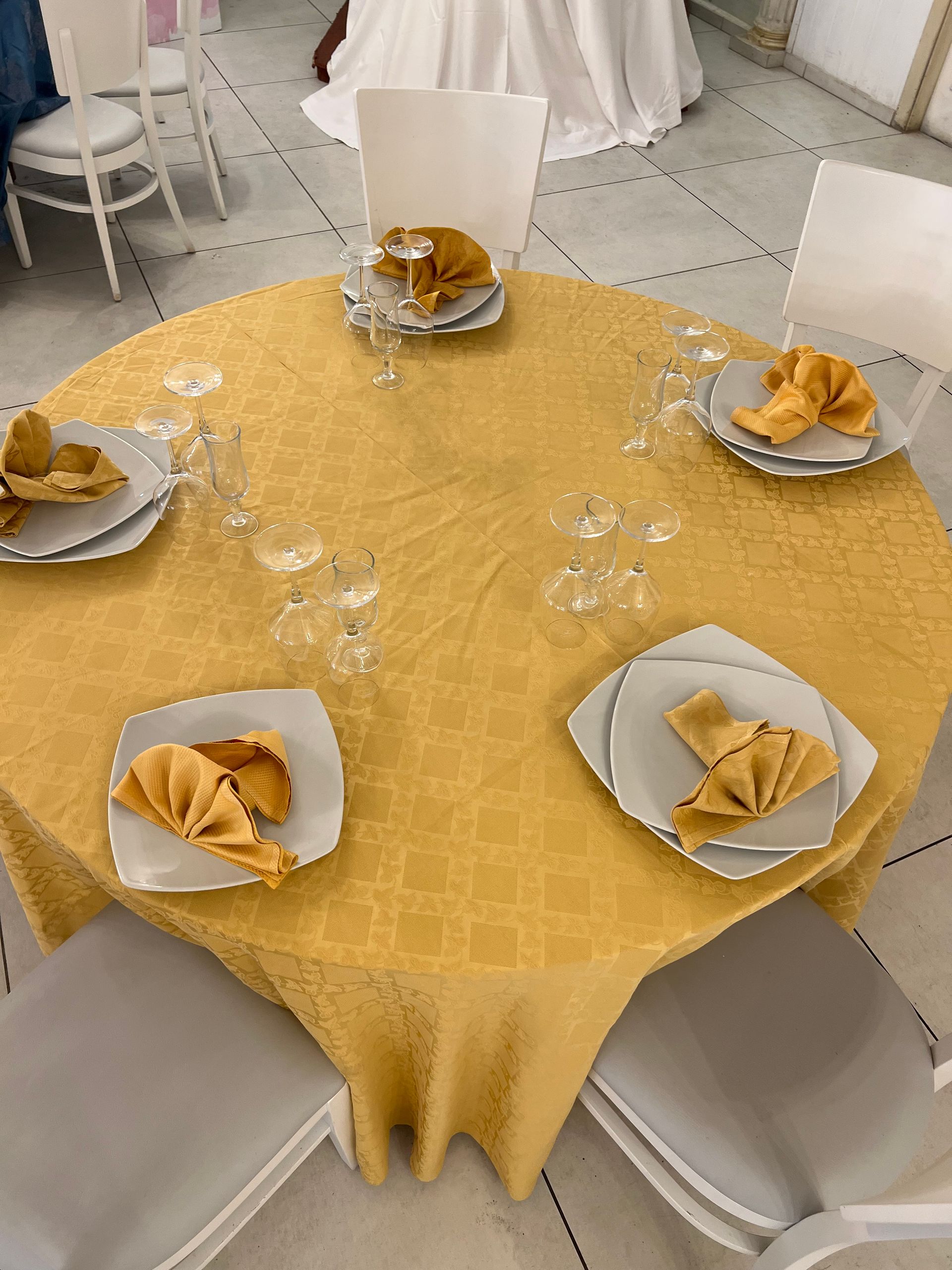 Tavola rotonda apparecchiata per un matrimonio, con tovaglia gialla, piatti bianchi, tovaglioli dorati e bicchieri.