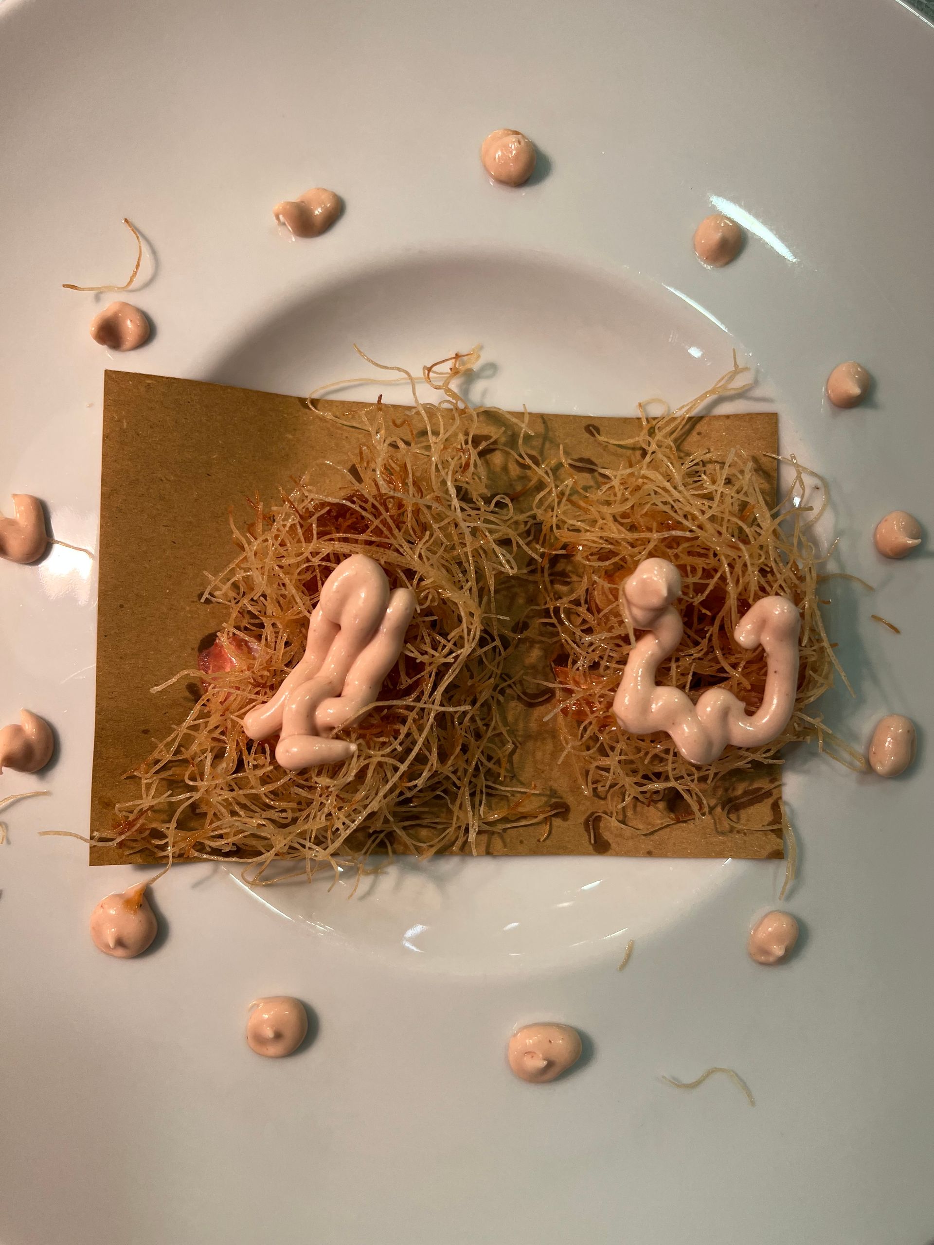 Due composizioni astratte di cibo su un piatto bianco con pois lungo i bordi. Ogni composizione ha una base marrone con fili dorati intrecciati e un vortice di salsa rosa.