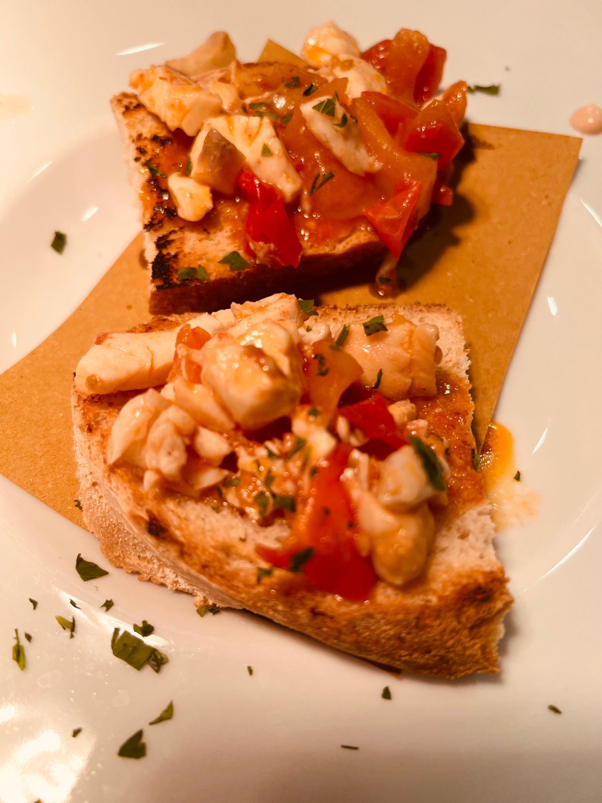 Due fette di bruschetta condite con frutti di mare, pomodori ed erbe aromatiche, su un piatto di carta e bianco.