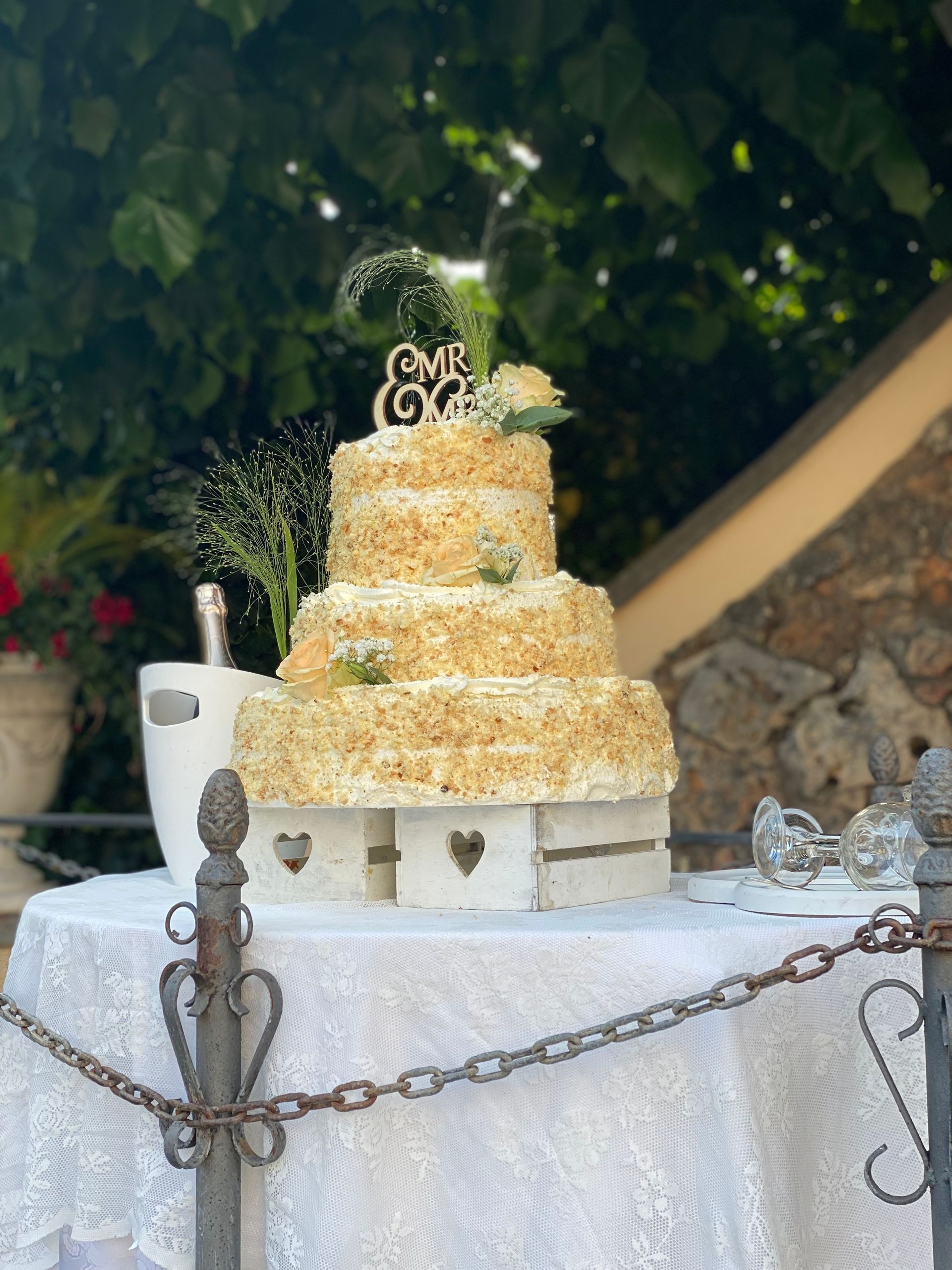 Torta nuziale a tre piani, decorata con fiori, su un tavolo bianco. Accanto, secchiello e bicchieri per champagne. Ambiente all'aperto.