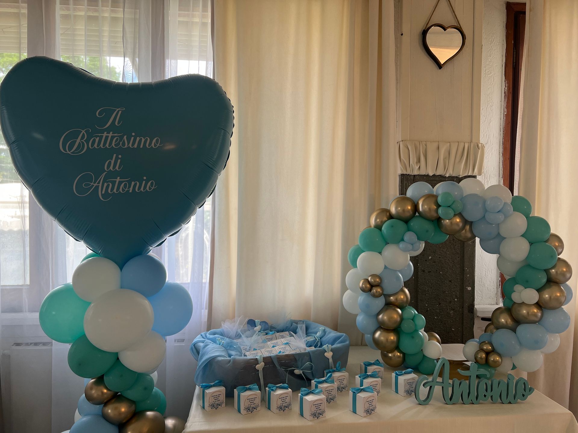 Decorazioni per il battesimo: palloncino a forma di cuore, corona di palloncini rotondi e tavolo dei regali con palloncini blu e oro.