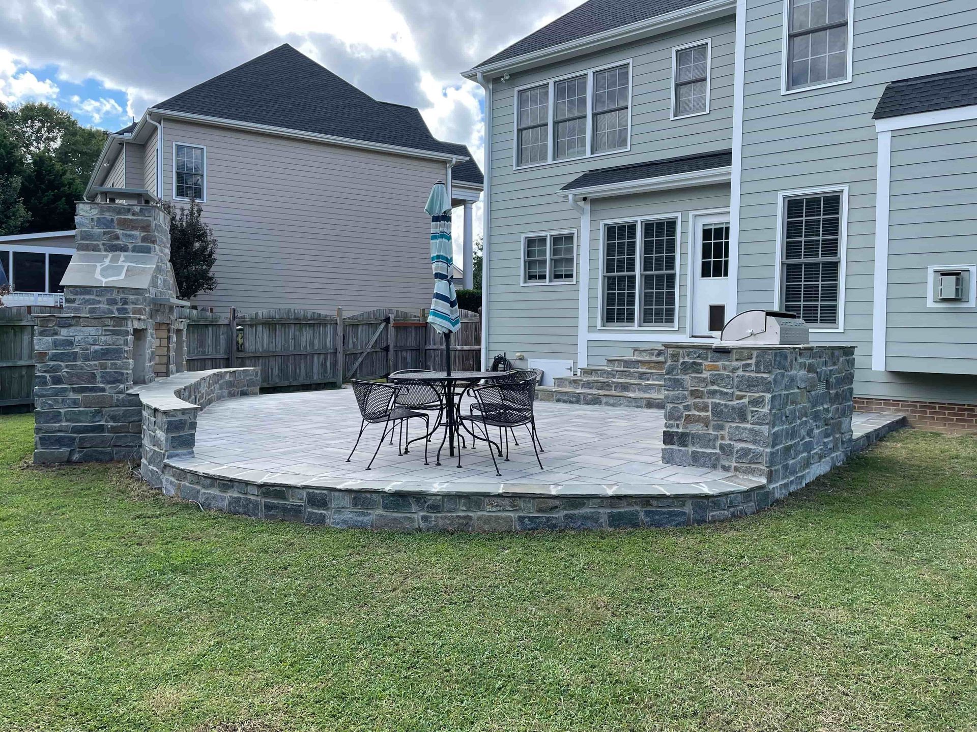 Back patio with stone retaining wall, patio table and chairs beside a gray house and grassy yard