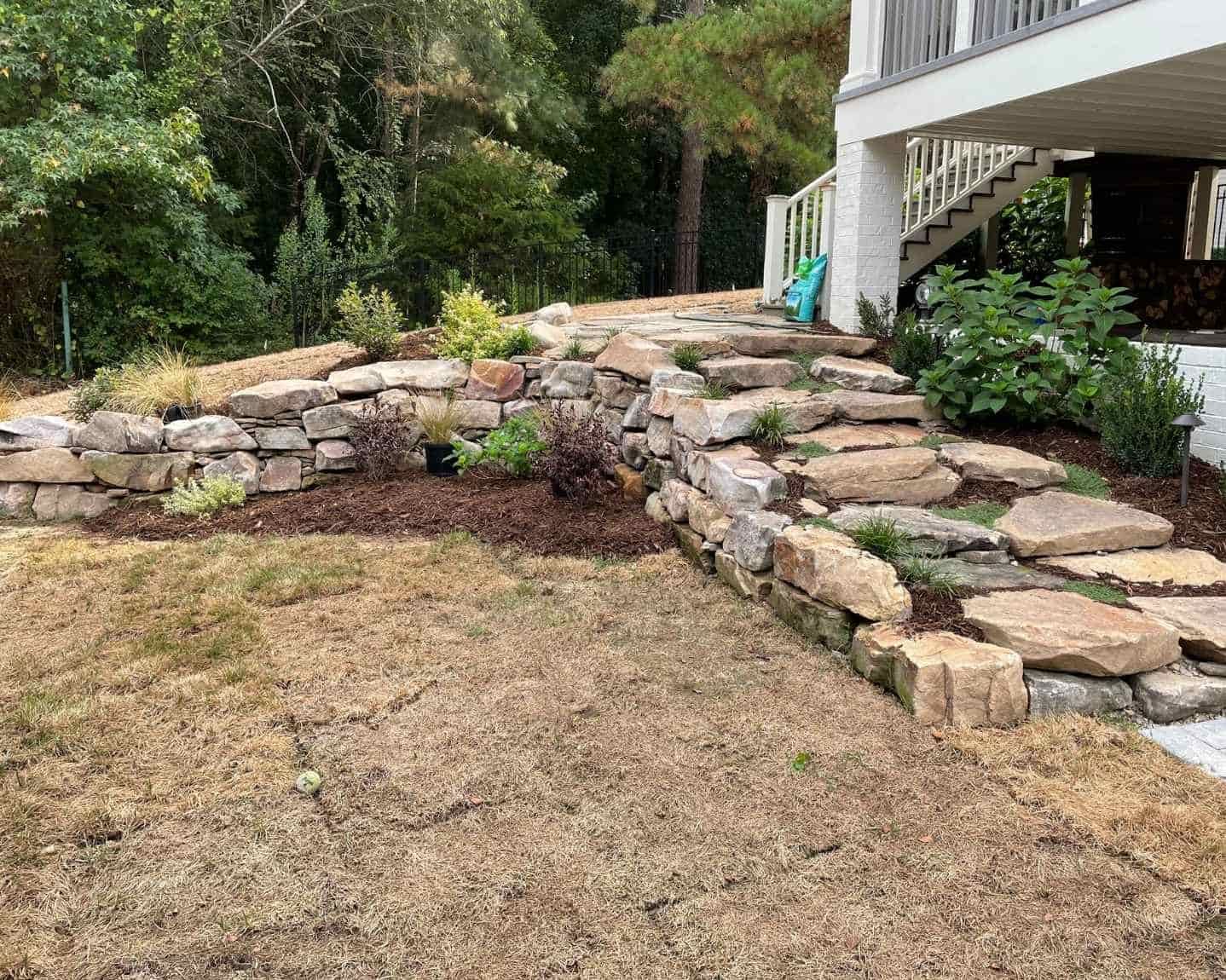 Stone retaining wall and steps leading to a porch beside a dry, brown lawn and trees