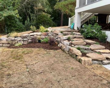Stone retaining wall and steps leading to a porch beside a dry, brown lawn and trees
