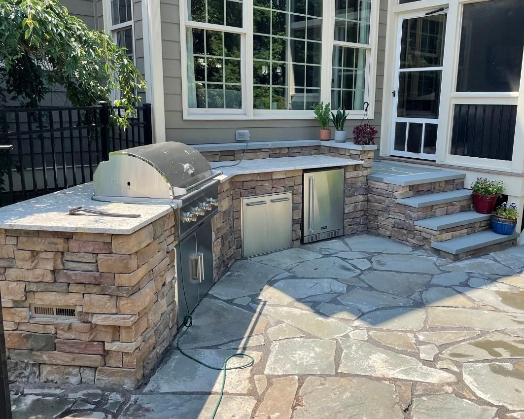 Stone outdoor kitchen and patio with built-in grill, cabinets, and a curved stone countertop beside a house.