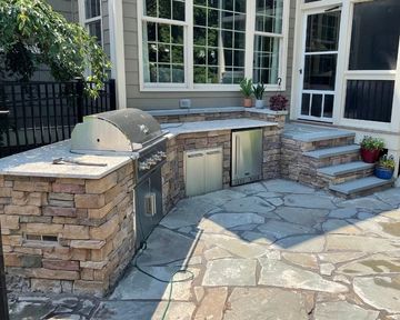 Stone outdoor kitchen and patio with built-in grill, cabinets, and a curved stone countertop beside a house.