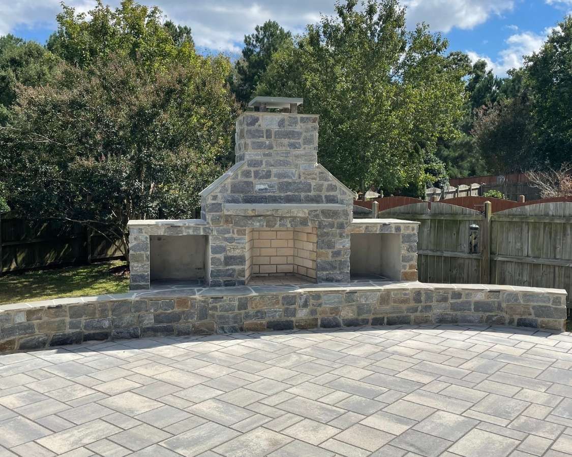 Stone outdoor fireplace with curved bench seating in a paved park area, framed by trees and blue sky