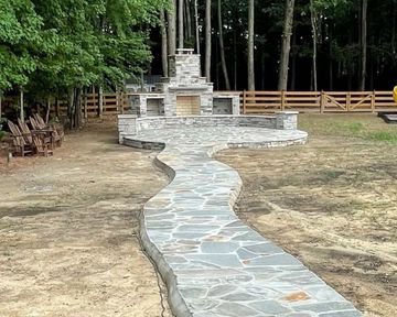 Curving stone pathway leading to a wooden fence and stone fireplace in a wooded yard
