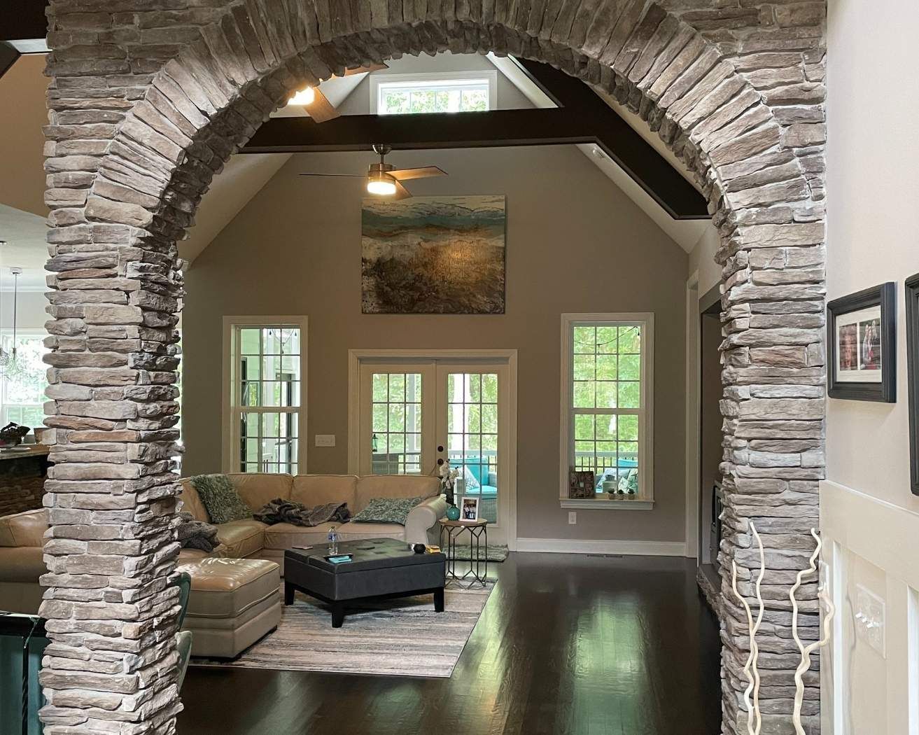 Stone archway opening to a bright living room with beige sofa, dark coffee table, and windows overlooking greenery