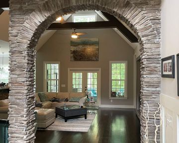 Stone archway opening to a bright living room with beige sofa, dark coffee table, and windows overlooking greenery