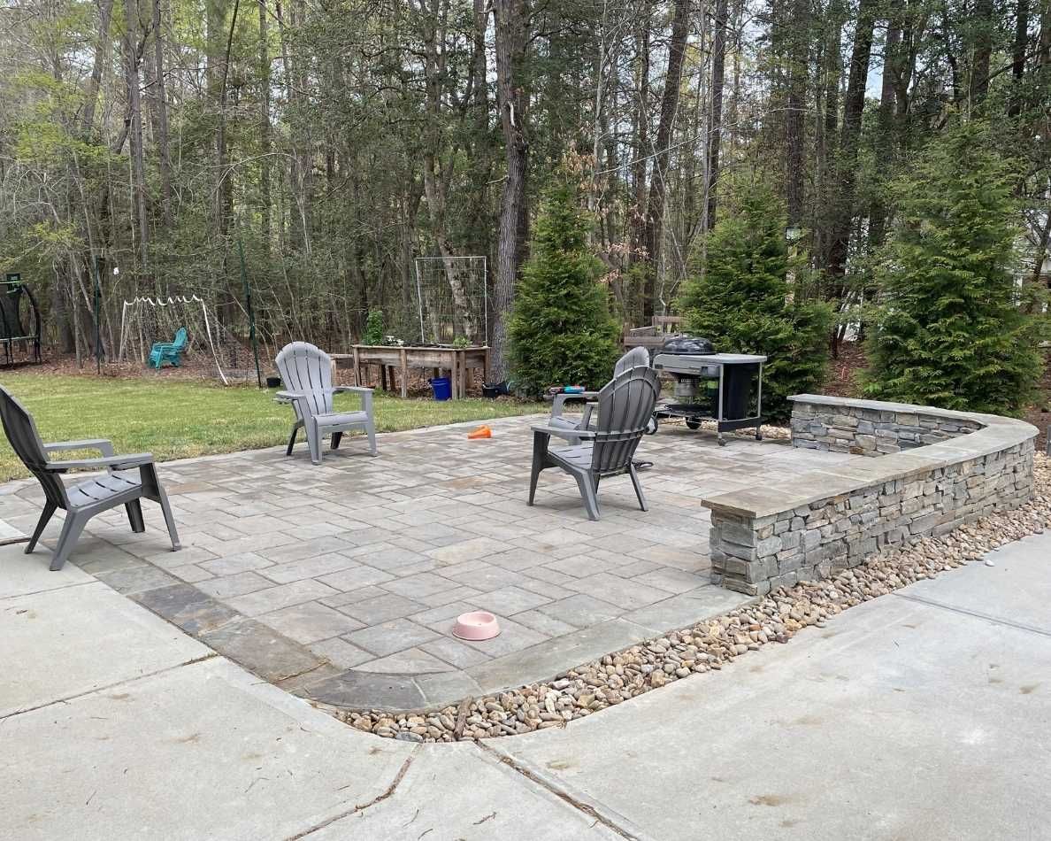 Stone patio with outdoor chairs and grill beside a wooded backyard and driveway