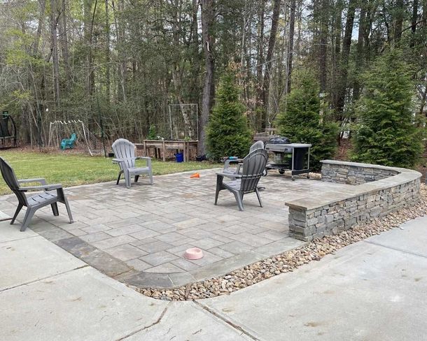 Stone patio with outdoor chairs and grill beside a wooded backyard and driveway