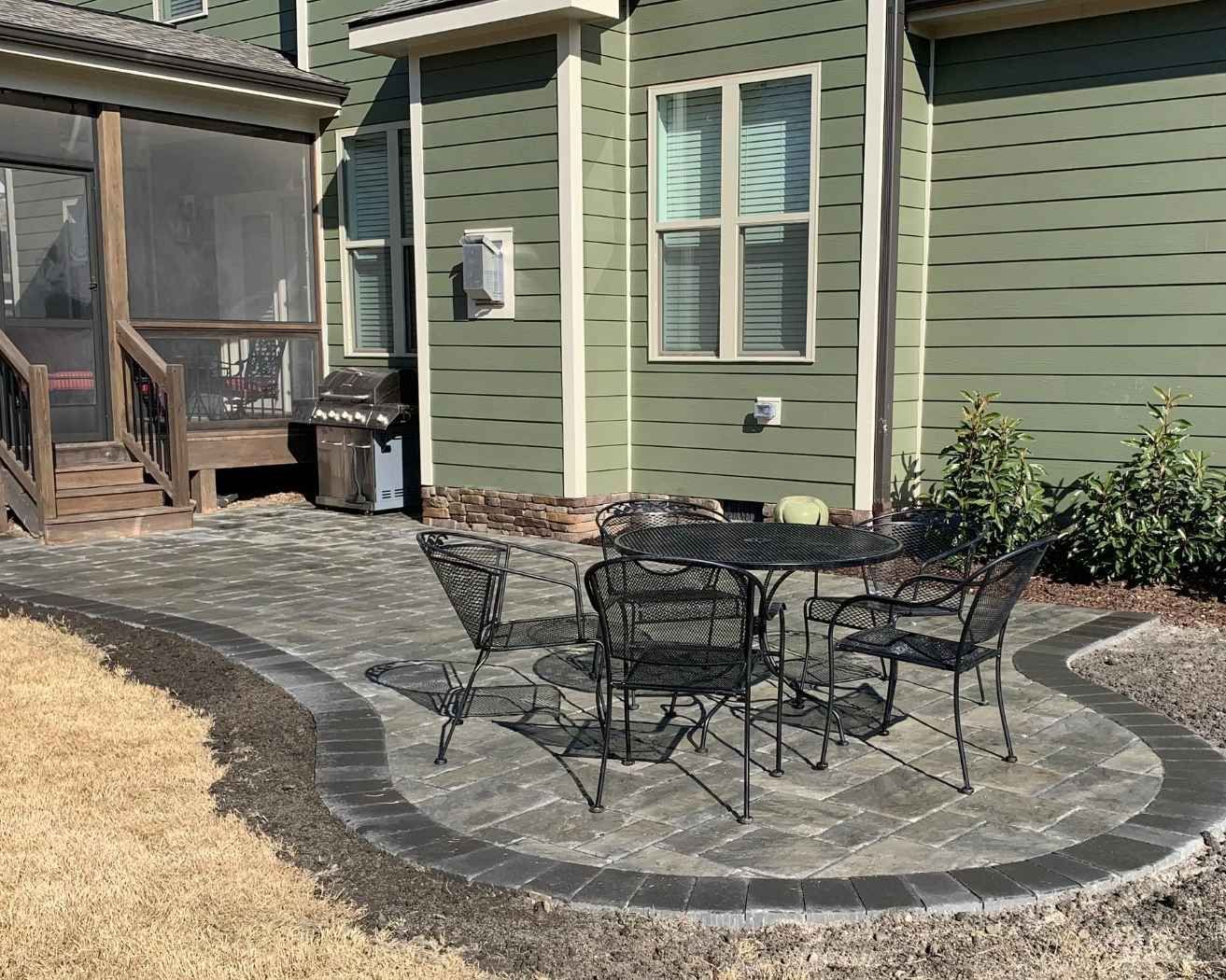 Backyard stone patio with black metal table and chairs beside a green house