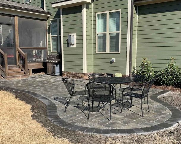 Backyard stone patio with black metal table and chairs beside a green house