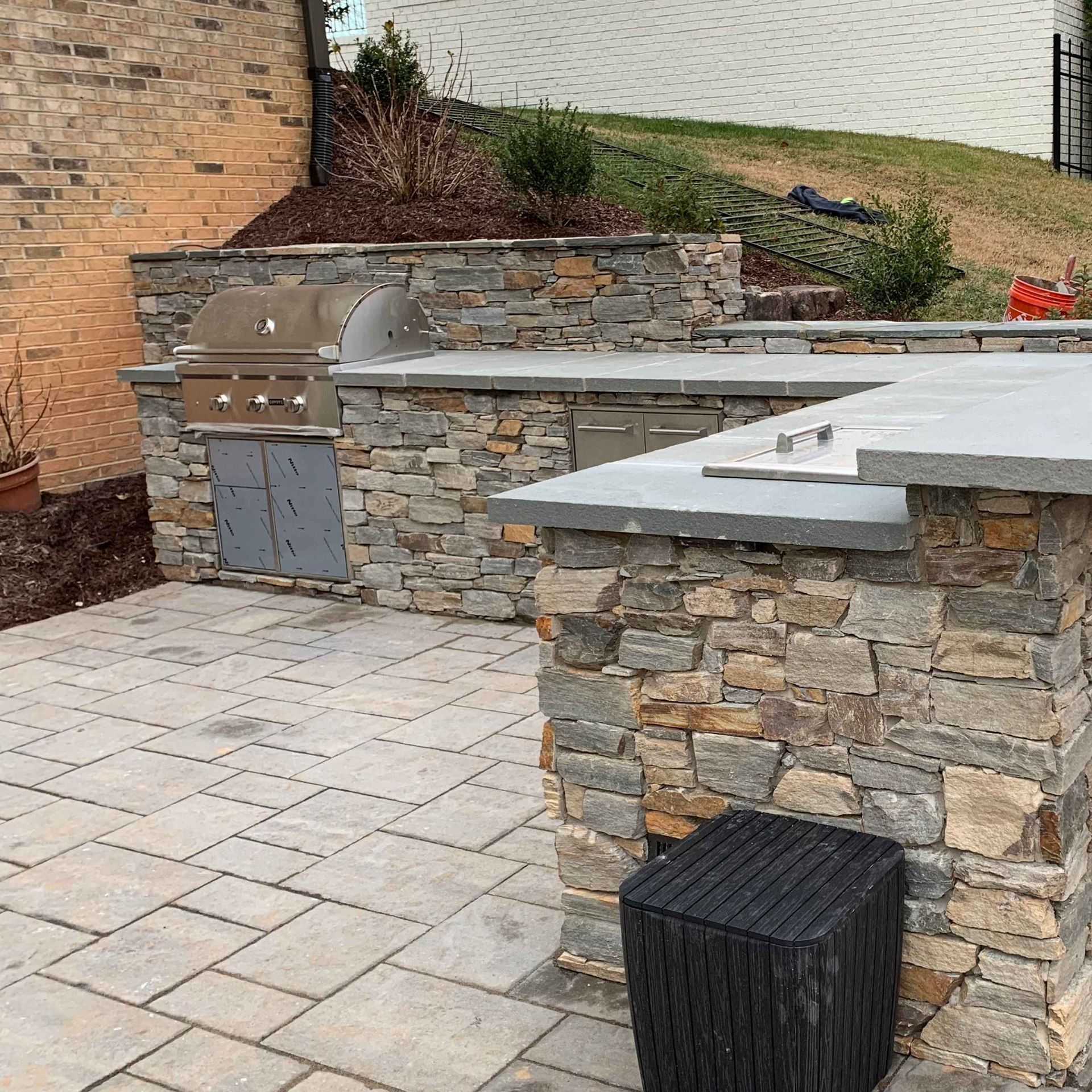 Stone outdoor kitchen with grill, countertop, and paver patio beside a brick house
