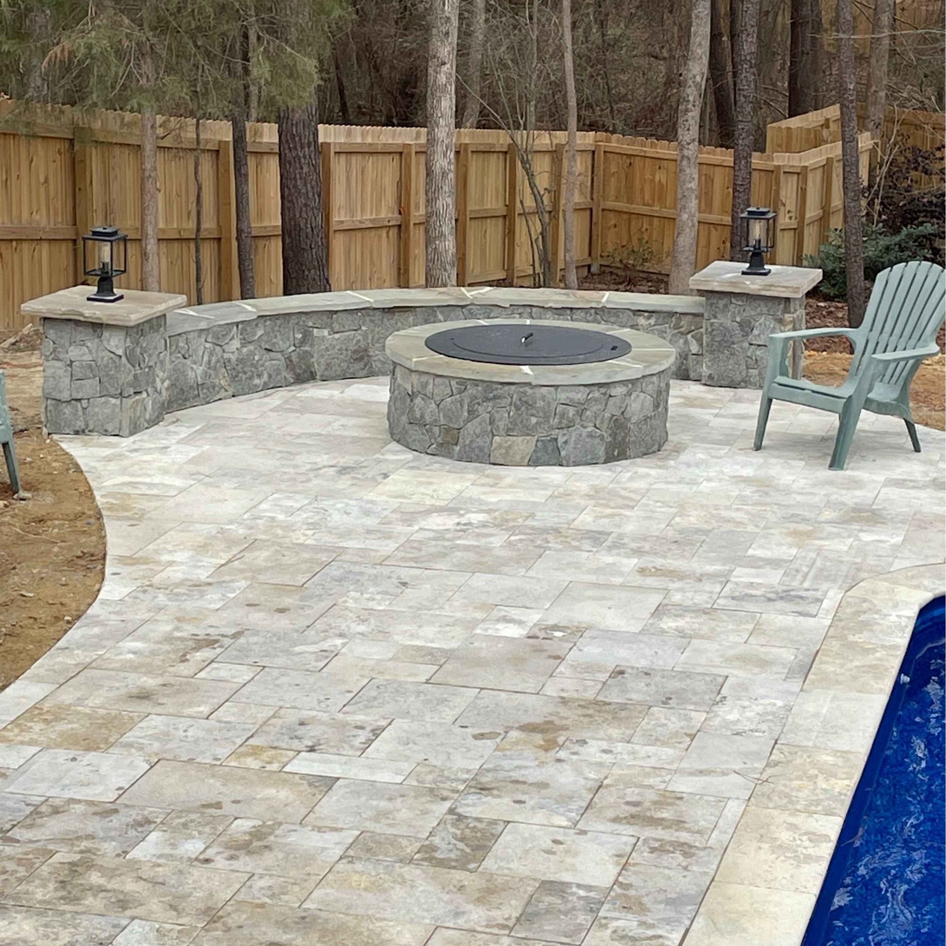 Stone patio with circular fire pit and seating area beside a pool, bordered by a wooden fence